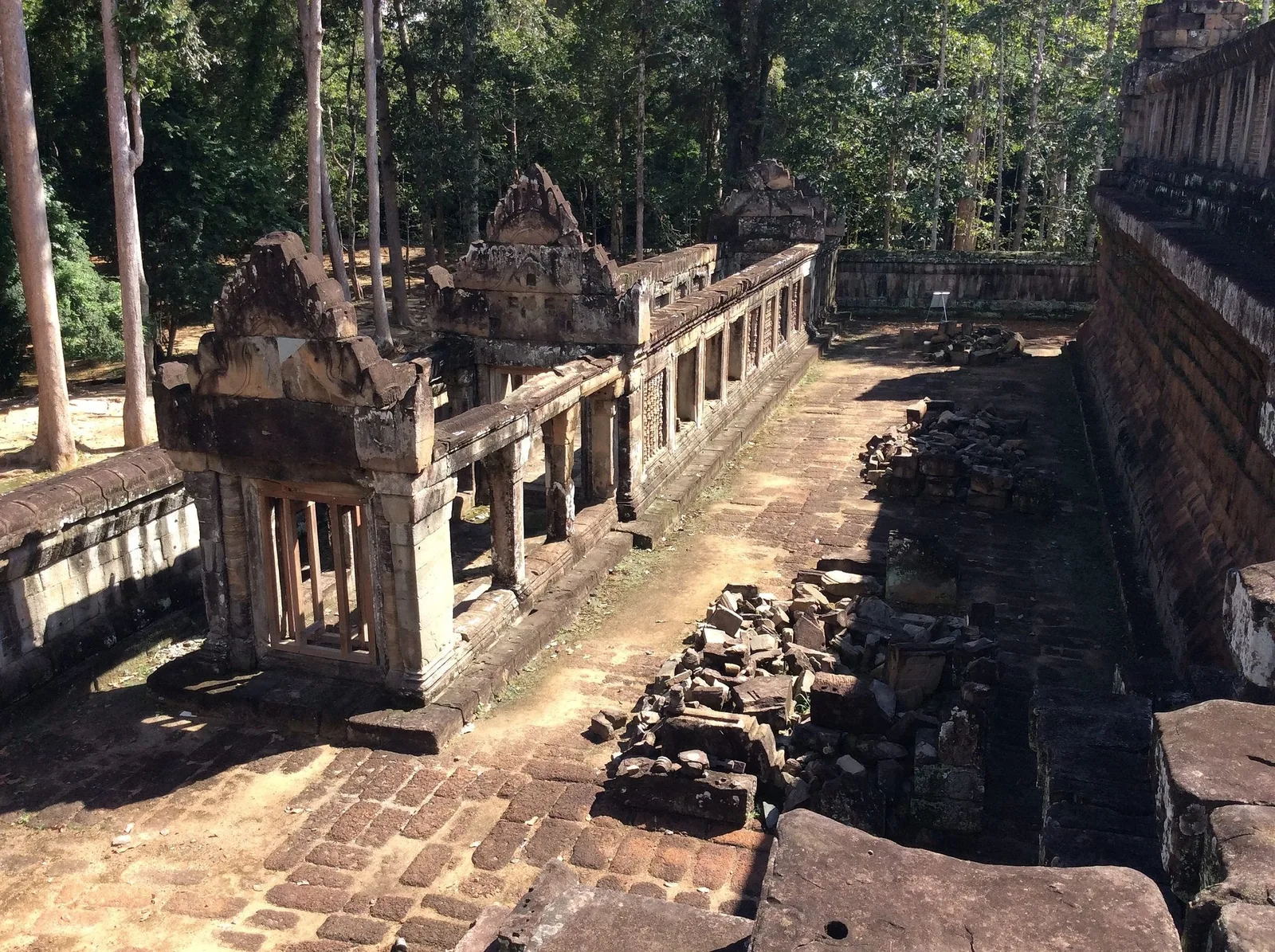 A ruined stone gallery with towers and scattered rubble stretches along the outer wall of an Angkor temple near Siem Reap.