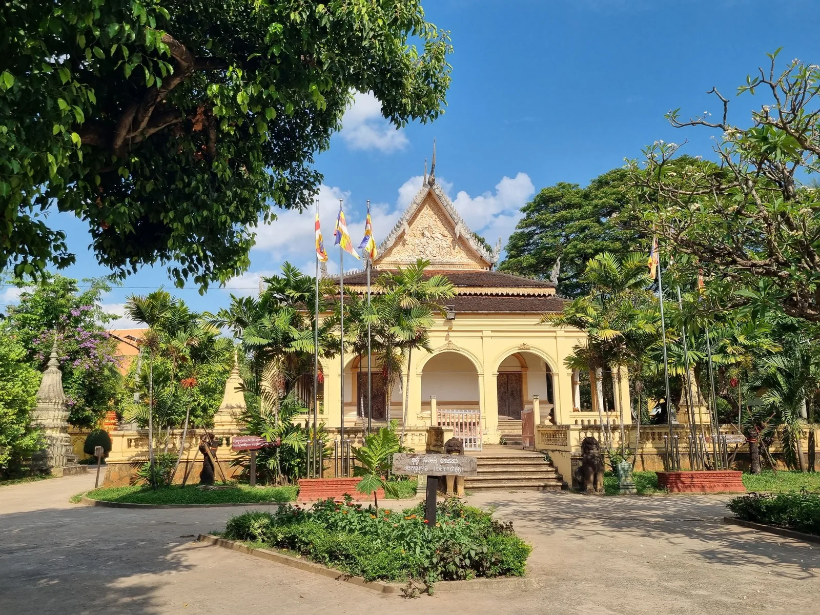Yellow pediment and columned porch of Wat Damnak temple surrounded by green trees under blue sky in Siem Reap