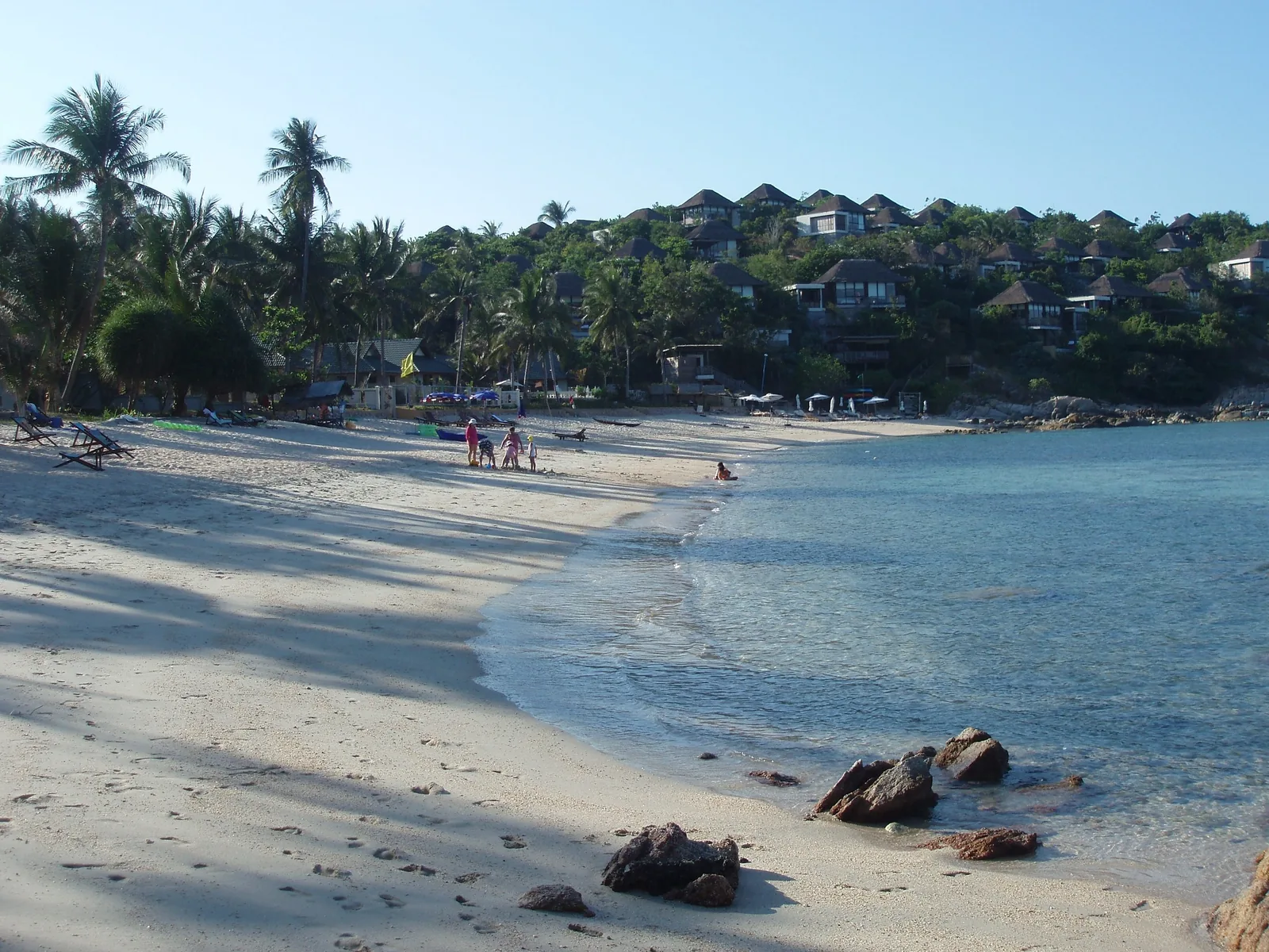 A curving white sand beach on Koh Samui with palm trees, hillside villas, and clear turquoise water.