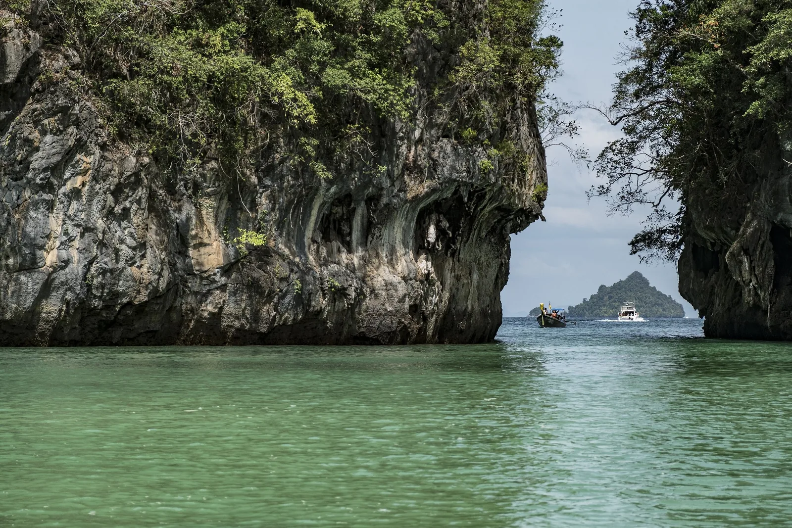 Limestone cliffs frame a turquoise lagoon and distant island near Krabi, with boats passing through the channel.