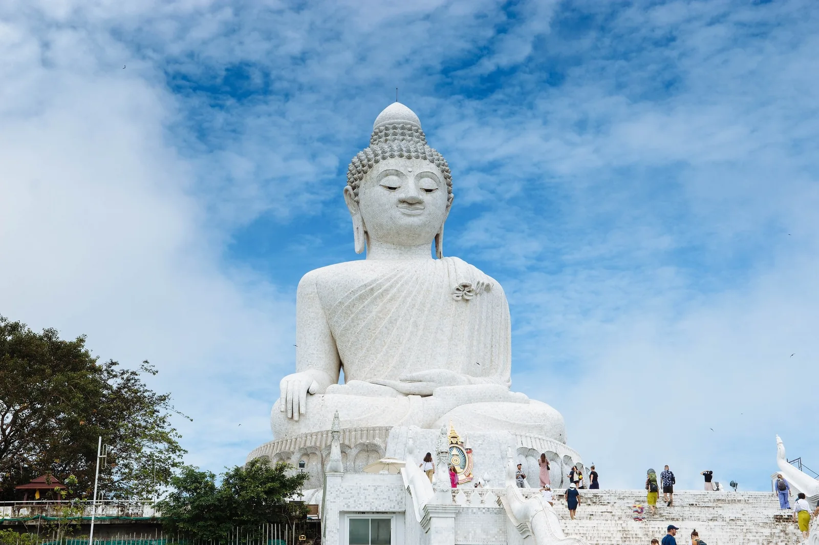 The Big Buddha's massive white marble statue rises against a blue sky above visitors climbing the stairs in Phuket.