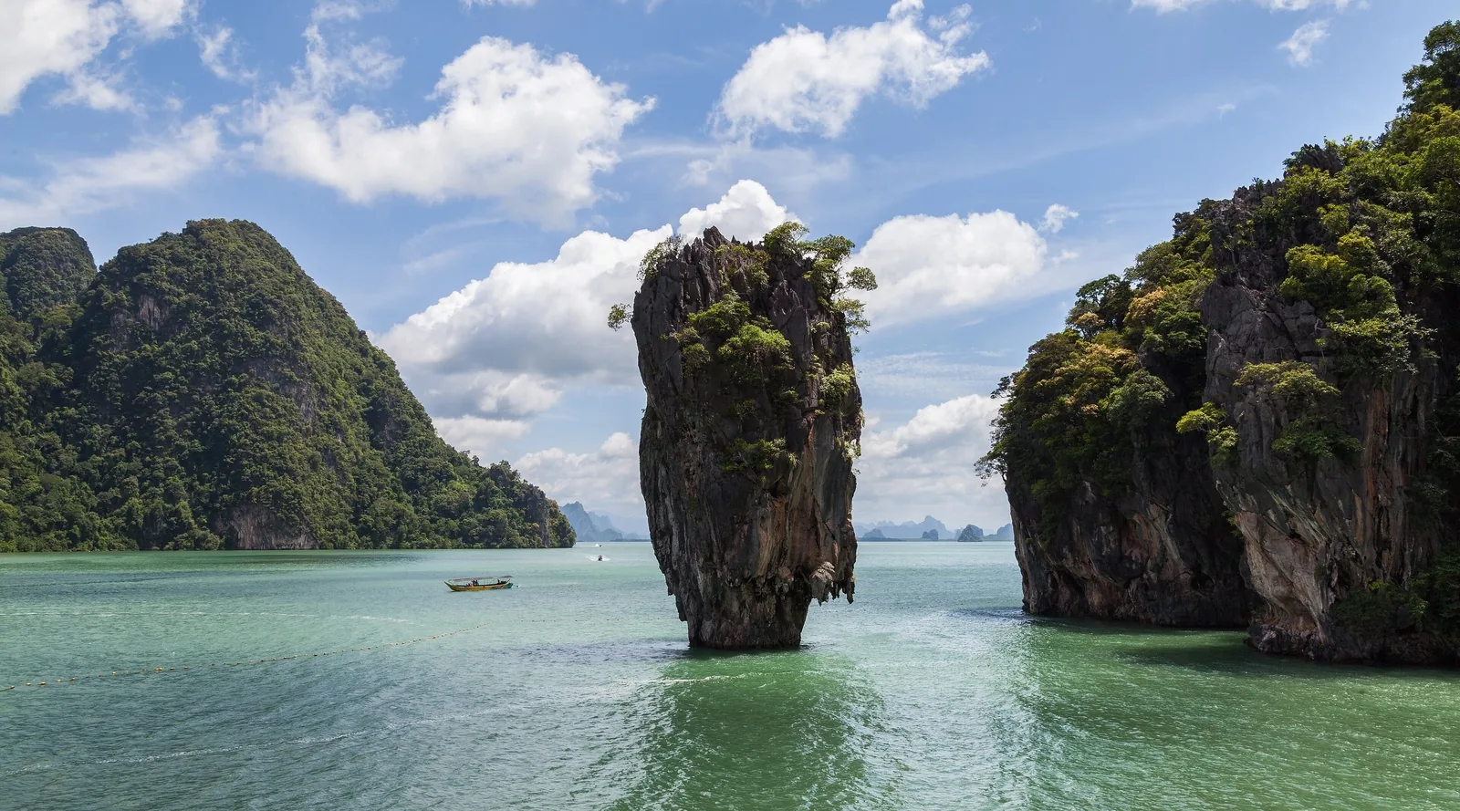 A striking limestone karst pillar rises from turquoise water near Phuket, Thailand, with a small boat nearby.